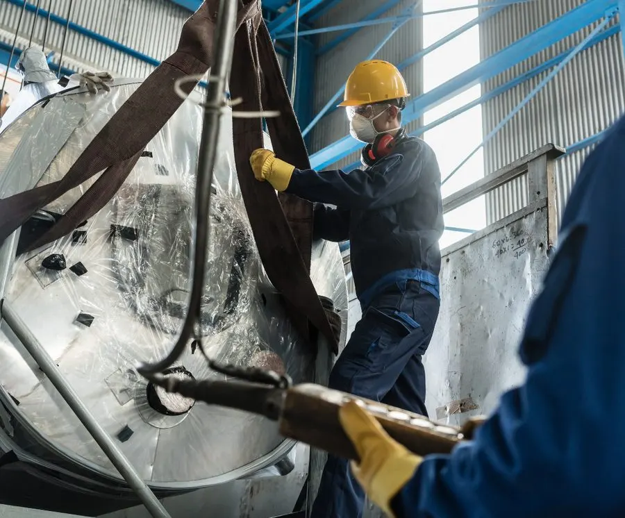 Profissionais com equipamentos de proteção içando um equipamento metálico em galpão industrial
